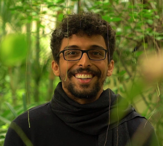 Foto de Bruno em um fundo de mata. Ele é jovem, moreno, usa óculos de grau, tem cabelos curtos encaracolados e usa barba