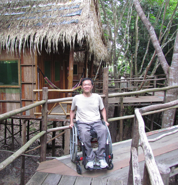 Ricardo, rapaz oriental vestindo camiseta branca e calça cinza está sentado em sua cadeira de rodas em uma passarela de madeira elevada que dá acesso a uma cabana que vemos ao fundo.