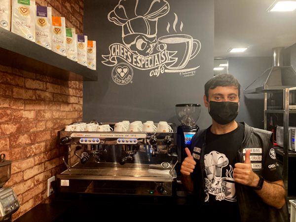 Na foto Fabricio posa ao lado de uma máquina profissional de café com o logo do café ao fundo.