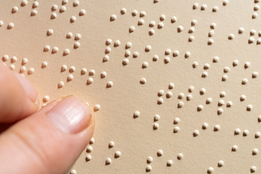 Foto em close de uma página escrita em braille. Um dedo está passando em cima dos pontos.