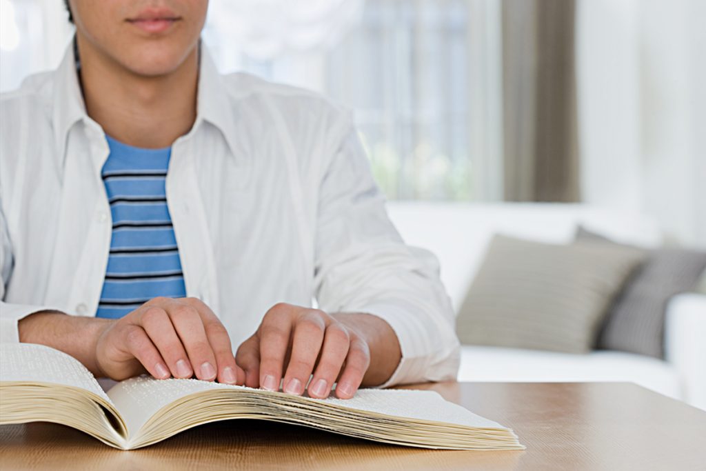 rapaz jovem, vestindo camisa branca aberta, sobre uma camiseta azul listrada, lendo um livro em braille
