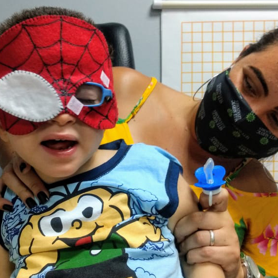 Criança vestindo uma camiseta do Cebolinha, está no colo da sua mãe usando uma máscara de feltro do Homem Aranha, com um dos olhos tapados pelo feltro.