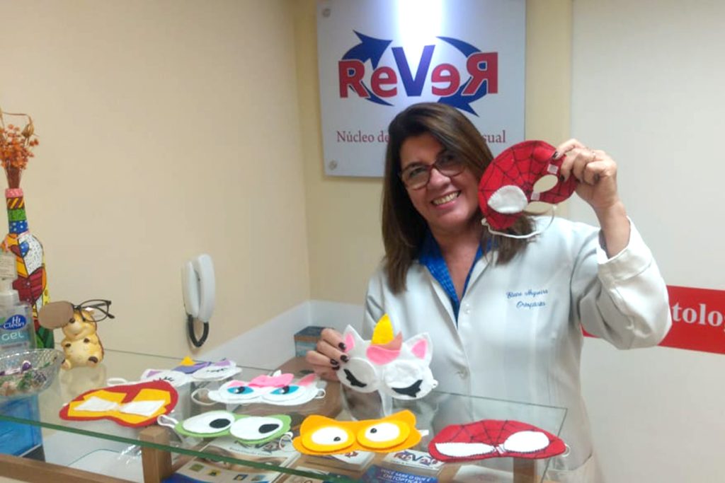 Eliane da clinica Rever, mostra as máscaras lúdicas feitas de feltro. Ela segura uma máscara do homem aranha em uma das mãos e uma máscara de unicórnio com a outra mão.