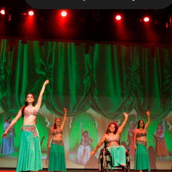 Foto de um palco com cortinas verdes de fundo e luzes vermelhas, tem 4 bailarinas vestidas com saia comprida verde e top de flores. As bailarinas estão de pé com um braço para cima e o outro para baixo. A bailarina do meio está sentada numa cadeira de rodas.
