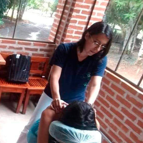 Foto de Andréia, moça de cabelos escuros, usando óculos de grau, camiseta azul marinho e calça branca, fazendo uma massagem em uma moça de cabelos escuros em uma cadeira de Quick Massage.