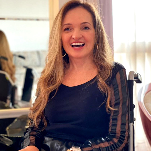 Foto de Beth Ribeiro, sorrindo sentada em uma cadeira de rodas. Ela é loira de cabelos compridos, usa uma blusa de manga comprida preta e brincos grandes de argola.