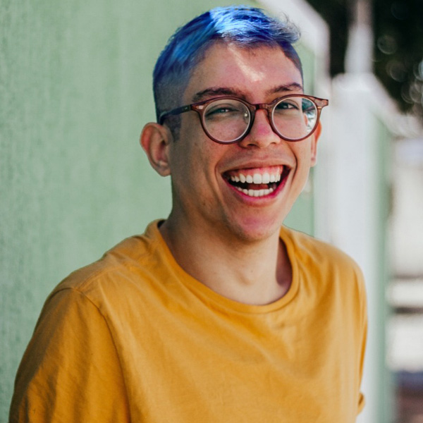 Foto do Ivan Baron sorrindo - jovem de cabelo curto azul, usando óculos de grau vestindo uma camiseta mostarda. Ele está em frente a um muro verde claro.