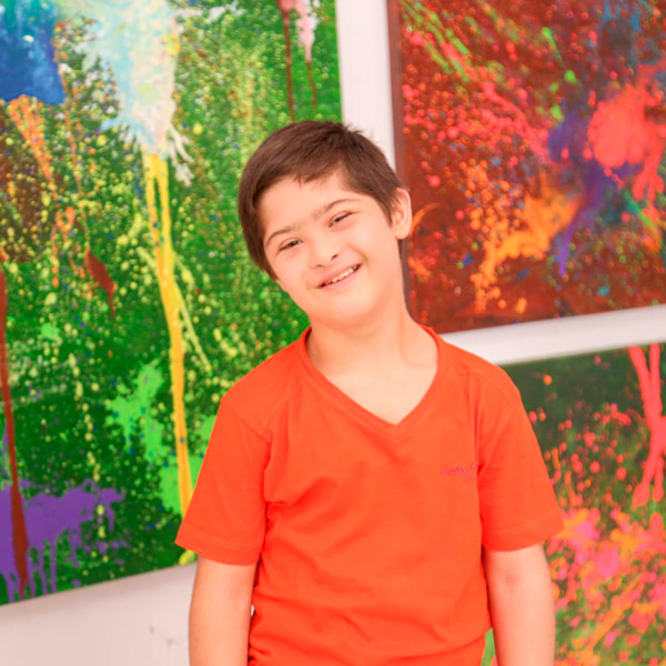 João Elyo, garoto de 12 anos, vestindo camiseta laranja, sorrindo em frente a uma parede com 3 quadros abstratos coloridos.
