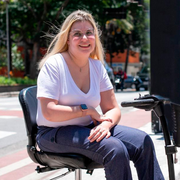 Foto de Nathalia Blagevitch sorrindo , sentada em uma cadeira motorizada na calçada de uma rua. Ela é loira de cabelos compridos, usa óculos de grau, uma camiseta branca e calça jeans.