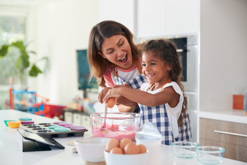 pra cego ver: foto da bancada de uma cozinha, onde vemos vários potes, ovos, uma forma de cupcakes com algumas forminhas coloridas e uma tijela onde uma menina de uns 5 anos está quebrando um ovo com a ajuda de sua mãe. Vemos também um tablet em cima do balcão, para onde a criança está olhando. Mãe e filha são morenas, usam um avental xadrez e estão sorrindo.