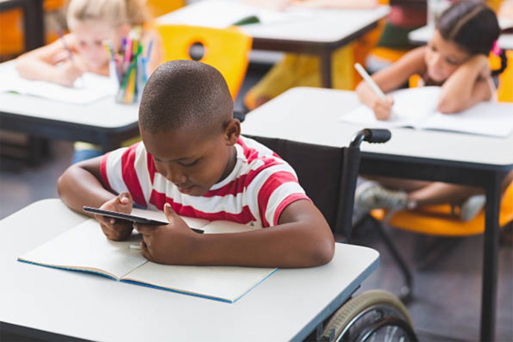 Em uma sala de aula, vemos um menino cadeirante usando um tablet enquanto os outros alunos estão usando papel e caneta.