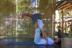 Bruno está deitado em um colchonete com as pernas para cima, sustentando uma aluna pelo quadril. Ela está com as pernas esticadas e o tronco para baixo.