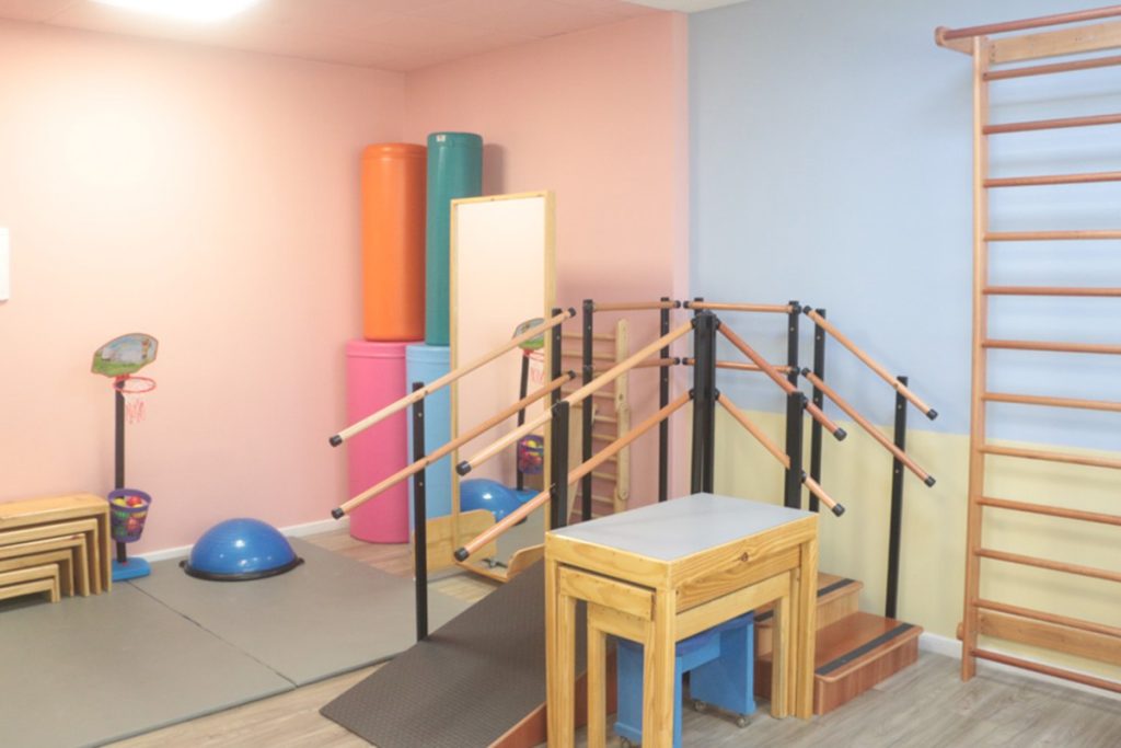 Pra cego ver: Foto do interior da clínica ProMover, onde vemos vários equipamentos de reabilitação e fisioterapia, como estepes, fitball, espaldar.. em uma sala com paredes rosa, azul e amarela.