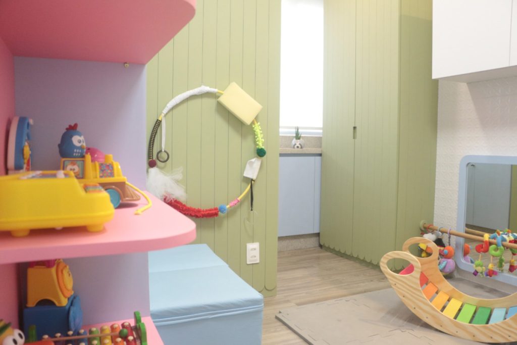 pra cego ver: foto de uma sala bem colorida, com armários verdes, fuffes azuis e prateleiras rosa, com diversos brinquedos infantis