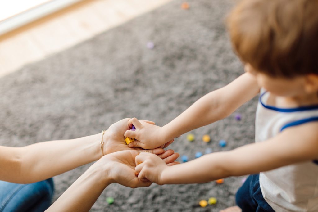 #pra cego ver: Criança está colocando nas mãos de um adulto diversas peças coloridas que vemos também espalhadas no carpete.