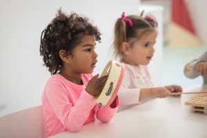 Foto de duas meninas pequenas sentadas em uma mesa, a primeira menina é bem morena e está vestindo camiseta rosa, com um pandeiro infantil na mão e a outra menina, loira de chiquinhas e camiseta clara está tocando xilofone.