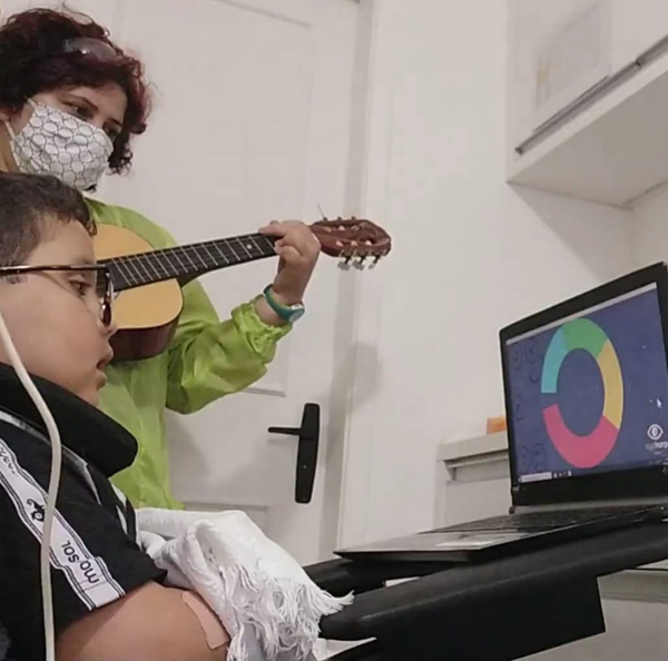 Foto de uma mulher tocando um violão ao lado de um menino que está em frente a tela de um notebook.