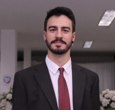 Foto de um rapaz jovem, moreno, com cabelos curtos e barba. Ele está vestindo um terno escuro com camisa branca e gravata vinho.