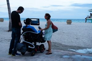Foto de uma praia, onde vemos um homem e uma mulher em pé ao lado de um homem em uma cadeira de rodas usando um dispositivo de Comunicação Alternativa acoplado em sua cadeira.