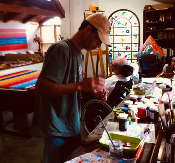 Foto de um atelie cheio de tintas e telas, onde em primeiro plano está um rapaz, manuseando tintas e ao fundo um senhos numa cadeira de rodas.