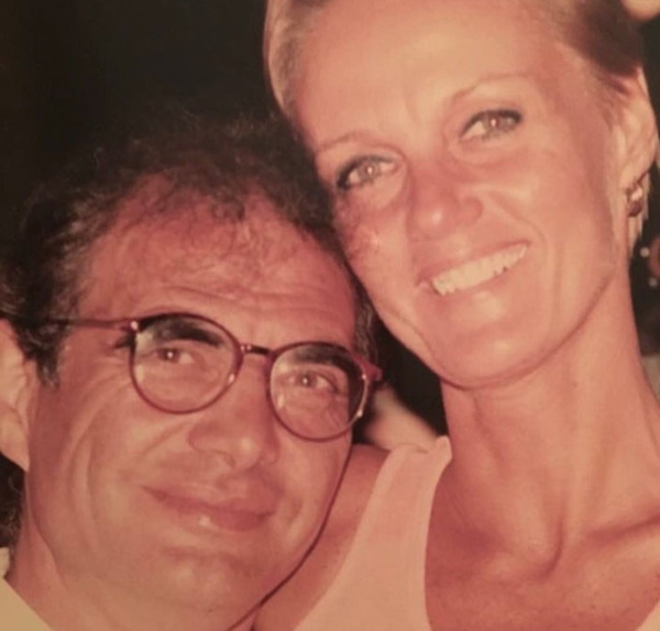 Foto em close de um casal. Ele é moreno e usa óculos e ela loira de olhos claros. Ambos estão sorrindo.