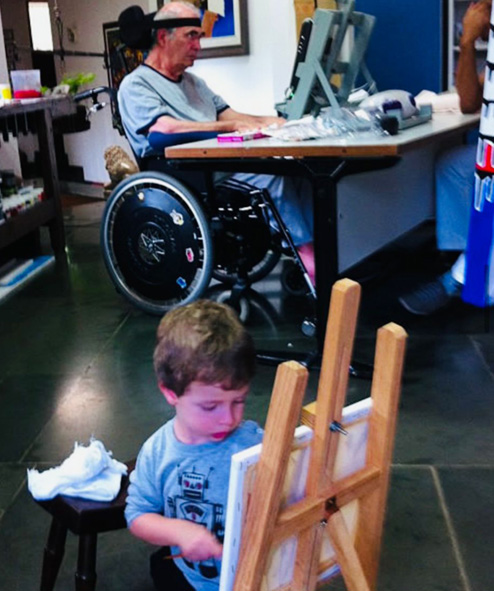 Foto de um atelier, e em primeiro plano um menino de uns 3 aninhos está pintando uma tela, ao fundo um senhor em uma cadeira de rodas com um suporte de computador em sua frente.