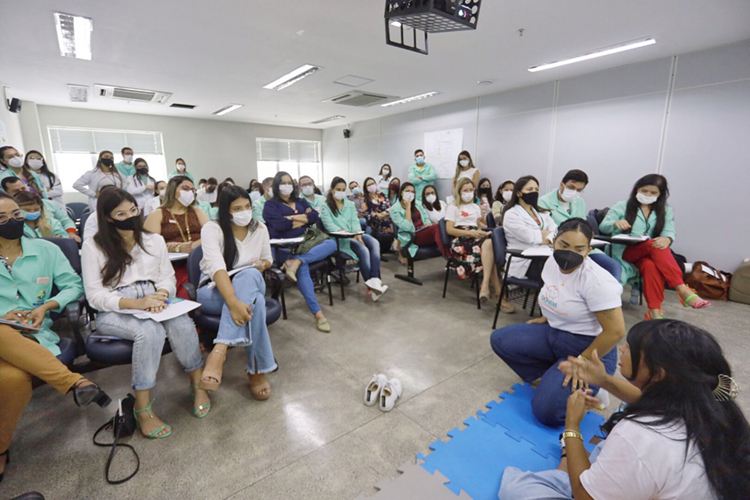 Foto de uma sala de aula cheia de pessoas com máscara, sentada em cadeiras e na frete duas mulheres estão sentadas no chão, explicando e gesticulando com as mãos.