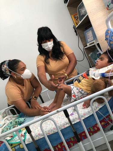 foto de uma cama infantil hospitalar onde vemos uma criança com traqueo, a mãe apoiada na cama e uma outra mulher em pé ao lado segurando o joelho da criança, explicando para a mãe.