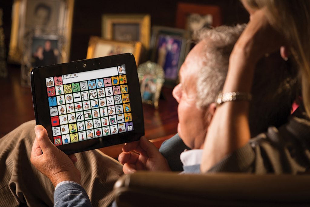 Um senhor com afasia está sentado em uma sala com um tablet de comunicação alternativa em sua mão. Atrás dele está uma mulher olhando para o tablet.