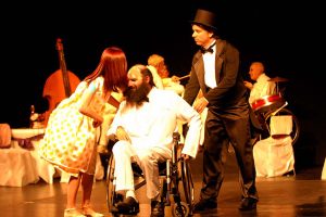 Na foto um cadeirante careca, com barba e vestindo um terno branco com gravata borboleta preta está em um palco de teatro com um homem de pé ao seu lado vestindo um smoking preto e usando uma cartola. Do outro lado está uma mulher abaixada cochichando em seu ouvido. Ela tem cabelos longos e usa um vestido branco de bolinhas. Ao fundo uma banda com 3 senhores.