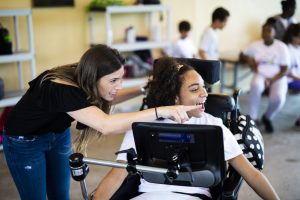 Em uma escola, uma menina morena sorrindo e olhando para o lado está em uma cadeira de rodas com um suporte que segura um tablet em sua frente. Ao lado dela está uma mulher de pé, apontando para o lado que a menina está olhando.