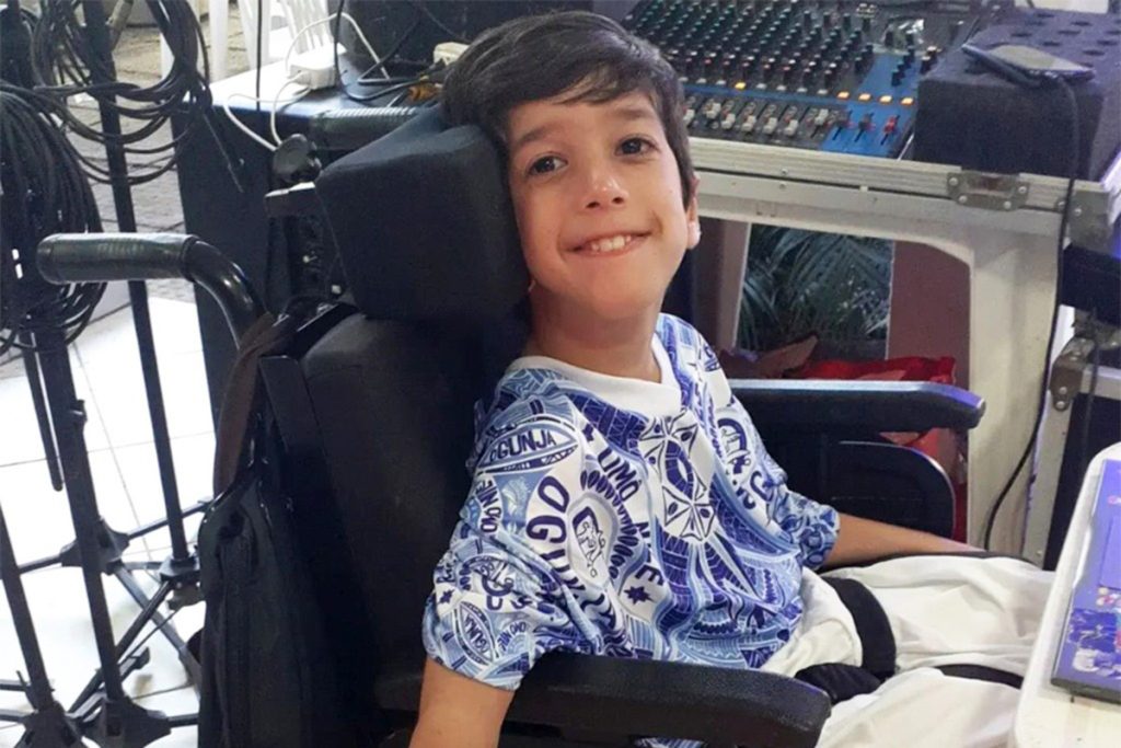 Garoto moreno, de 10 anos, sorrindo, vestindo uma camiseta estampada em tons azuis, sentado em uma cadeira de rodas, com uma mesa de som e vários cabos ao seu lado.