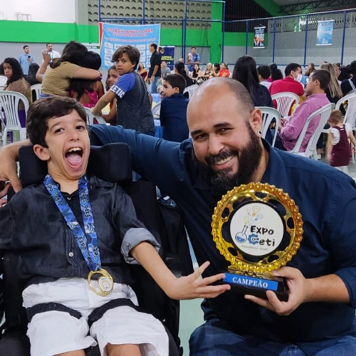 Menino de 10 anos, com cabelos escuros, vestindo camisa azul, bermuda branca e com uma medalha no pescoço, sentado em uma cadeira de rodas está ao lado de um rapaz careca e com barba, também usando uma camisa azul e sorrindo, que está segurando um troféu. Ao fundo, diversas pessoas sentadas em cadeiras de plástico em uma escola.