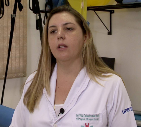 Foto de uma mulher jovem com cabelos longos e loiros. Ela veste um jaleco branco e está em uma sala de terapia.