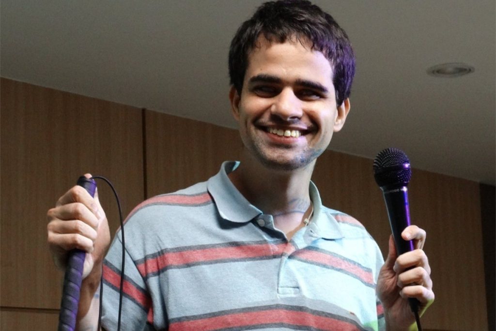 Rapaz com cabelos escuros, sorrindo, está vestindo uma camiseta polo azul com listras vermelhas e está segurando um microfone com uma das mãos e na outra uma bengala.