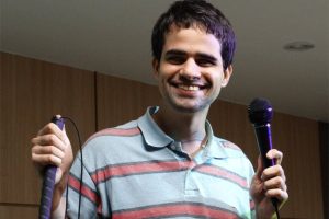Rapaz com cabelos escuros, sorrindo, está vestindo uma camiseta polo azul com listras vermelhas e está segurando um microfone com uma das mãos e na outra uma bengala.