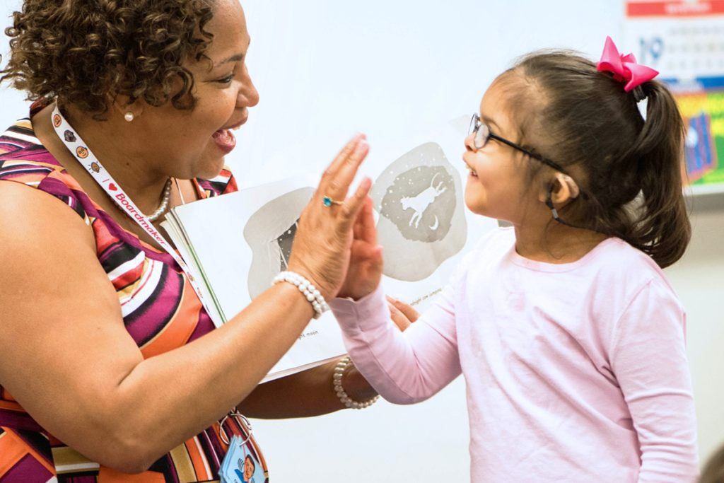 Foto de uma senhora morena com cabelos curtos enrolados, usando um vestido estampado e um cordão de comunicação Alternativa e segurando um livro com figuras aberto, está sorrindo olhando para uma menininha e batendo em sua mão em forma de "comemoração". A menininha morena usando óculos, cabelos presos com um laço rosa e camiseta rosa, também está sorrindo olhando para a senhora.