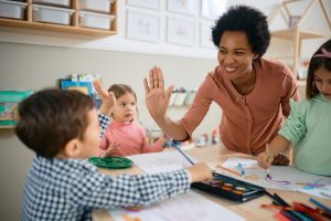Foto de uma uma mesa de sala de aula infantil com duas meninas e um menino de uns 3 anos sentados pintando com aquarela. Uma professora está de pé ao lado sorrindo e dando um comprimento ao menino, batendo a palma da mão dela na dele.