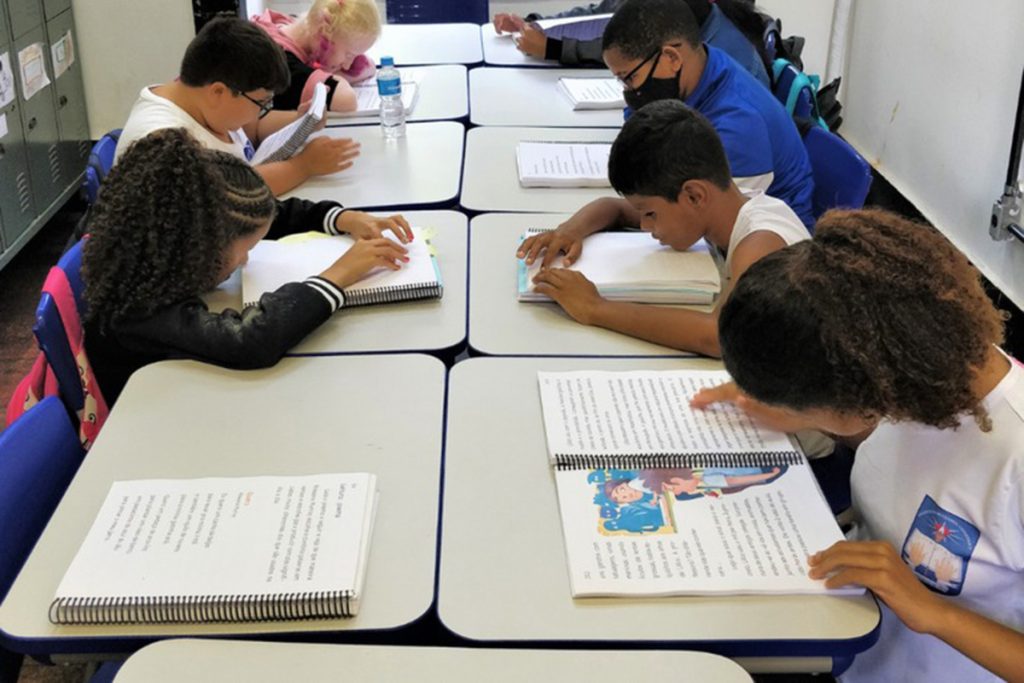 Foto de 8 crianças sentadas em carteiras escolares, lendo livros pelo método Braille. Todas estão vestindo uniformes em tons de azul e branco.
