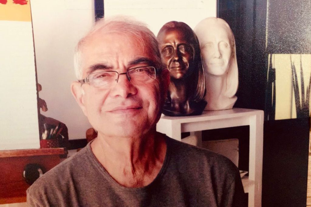 Foto de um senhor grisalho usando óculos e camiseta marrom. Ele está em frente a dois bustos (esculturas) de uma mulher. Um feito de bronze e o outro de gesso.
