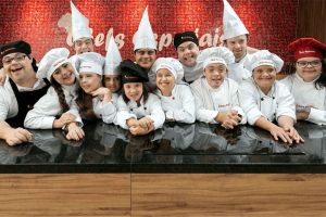Foto de um balcão de cozinha com o fundo vermelho com o letreiro: Chefe Especiais. Encostados atrás do balcão estão 13 jovens com Síndrome de Down usando domas e chapéu de chef. Todos estão sorrindo.