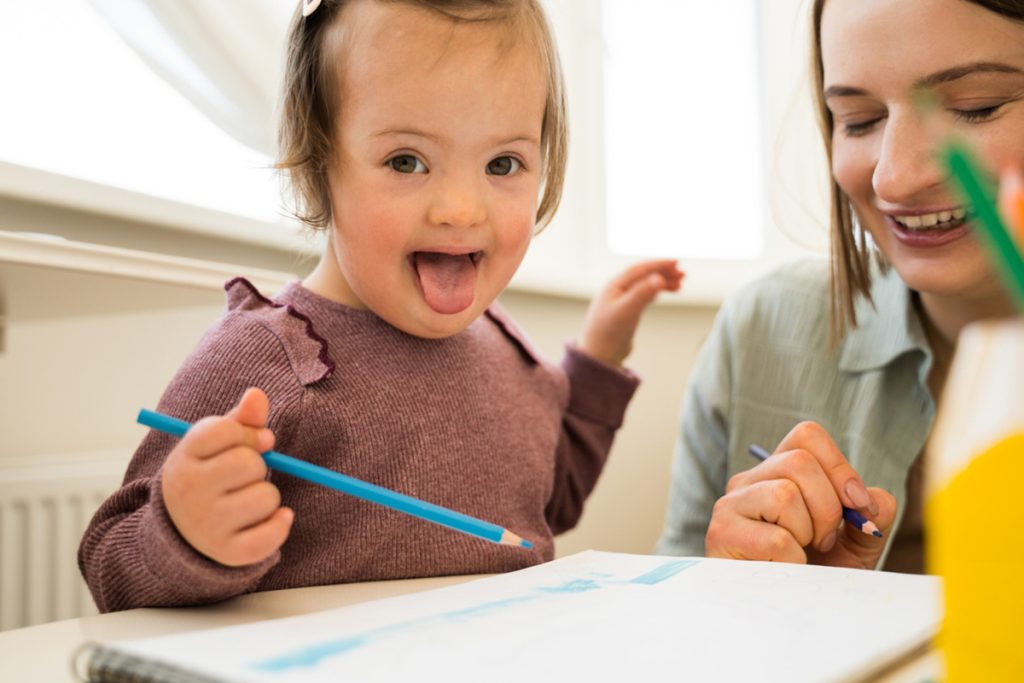 Uma menina com síndrome de down de uns 3 anos vestindo uma blusa marrom com a língua de fora, está sentada em uma mesa de escola segurando um lápis azul e em sua frente um caderno de desenho . Ao seu lado uma professora de camisa verde também segura um lápis sorrindo e olhando para o caderno.