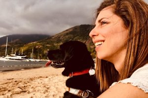 Foto em close de Mellina com sua cadela Hilary em uma praia com barcos ao fundo. Mellina é loira de olhos claros e Hilary é uma labradora preta. Ambas estão sorrindo.