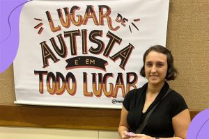Foto da Alice, uma garota jovem de cabelos curtos vestindo uma blusa preta, sorrindo em frente a um banner que está escrito: Lugar de autista é em todo lugar.