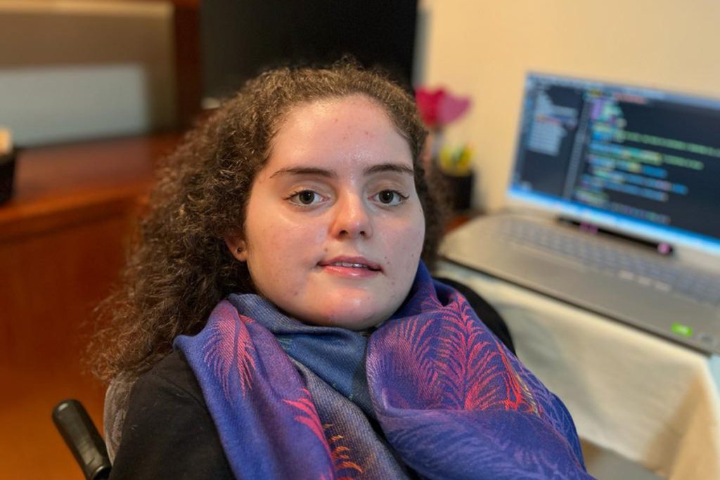 Foto da Julia sentada em uma cadeira em seu escritório. Ao fundo o computador com o rastreador ocular. Julia é jovem, com cabelos enrolados presos, tem olhos castanho claros e veste camiseta preta e uma echarpe lilás ao redor do pescoço.