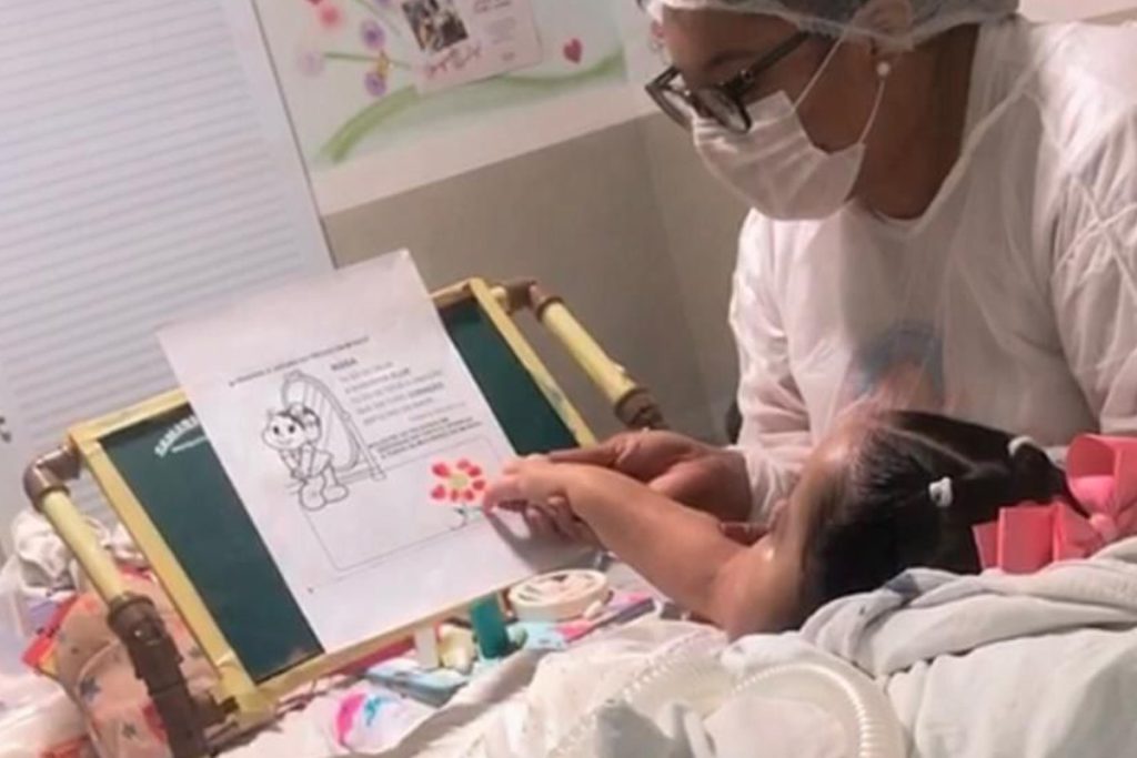 Foto de uma garotinha em uma cama, com uma prancheta em sua frente com uma atividade escolar. Ela está pintando com os dedos, com ajuda de uma pedagoga que está ao seu lado.