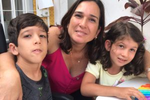 Foto de uma mulher de pele clara, cabelos escuros lisos na altura do ombro, usando uma regata rosa, abraçando seus dois filhos. Na esquerda o filho de uns 11 anos, cabelos escuros curtos, vestindo camiseta cinza, e na direita a filha de uns 8 anos, cabelos castanhos encaracolados na altura dos ombros vestindo uma camiseta amarela. Todos estão sorrindo.
