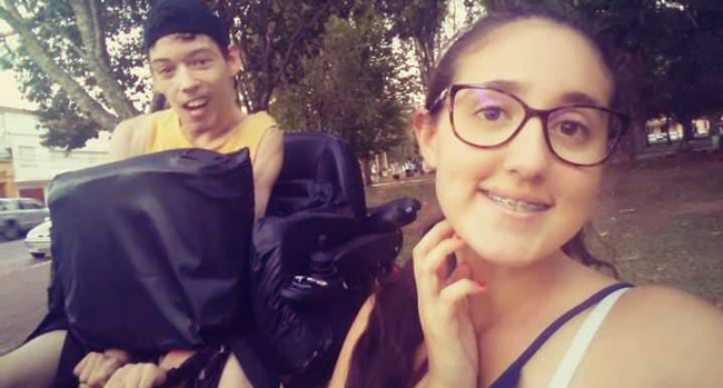 Foto de um rapaz com boné pretoao contrário, vestindo regata amarela em uma cadeira de rodas e ao seu lado uma jovem de cabelos presos usando óculos de gral e aparelho nos dentes.