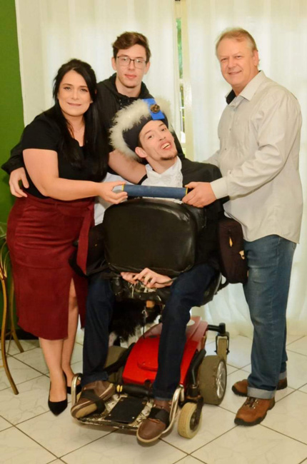 Foto de uma familia. A mãe no lado esquerdo de pé, vestindo uma blusa preta, saia vermelha e sapatos de salto, o pai no lado direito vestindo camisa clara e calça jeans, o filho mais velho ao centro em uma cadeira de rodas usando beca e o filho mais novo atrás usando óculos. O pai e a mãe estão segurando um tubo de diploma em frente ao filho cadeirante.