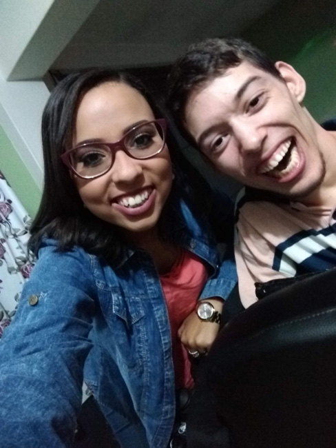 Foto de uma jovem morena com cabelos escuros na altura do ombro, usando óculos de grau e vestindo uma camisa jeans, ao seu lado um jovem sorrindo vestindo camiseta listrada.